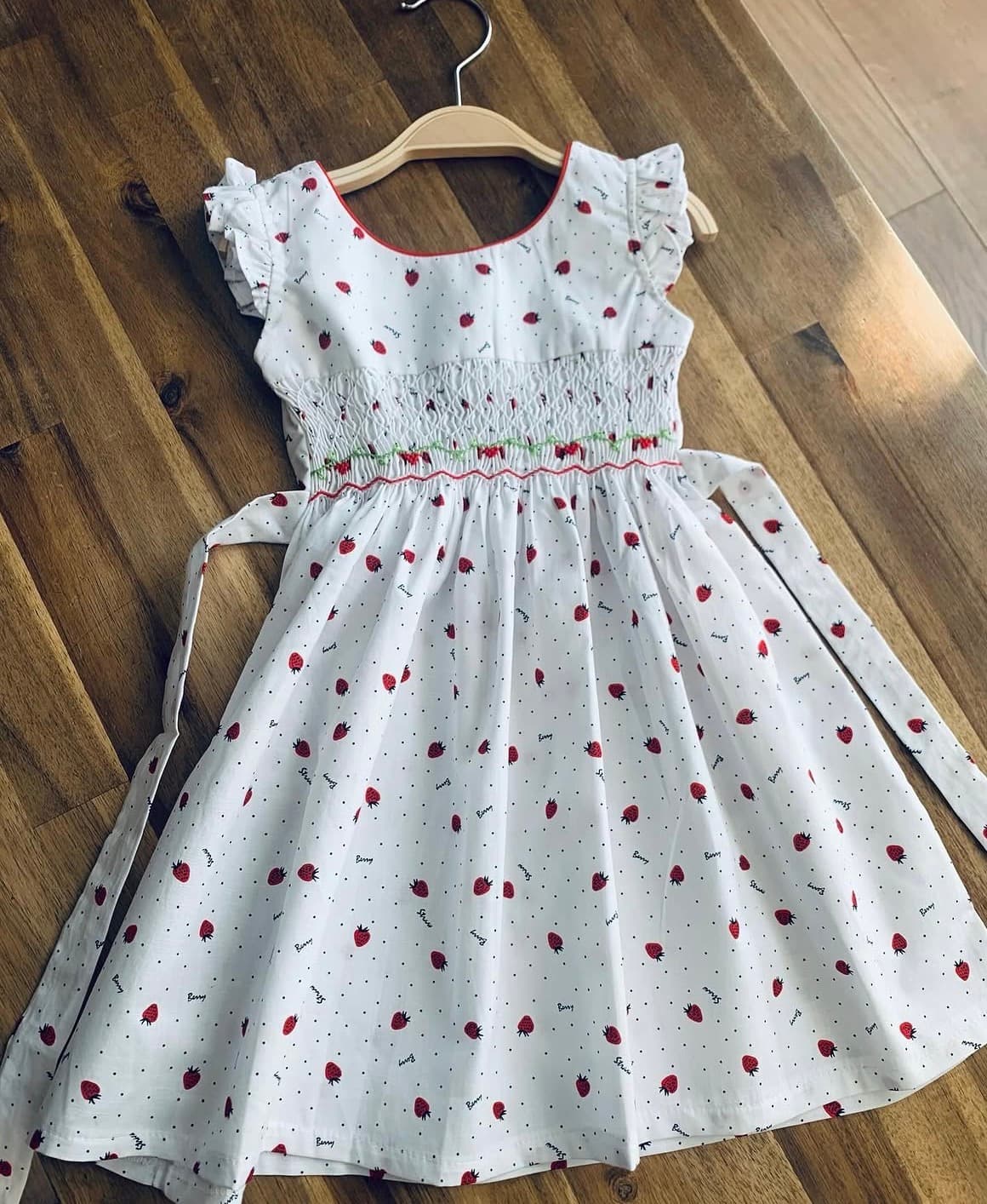 White dress with strawberry print, smocked bodice, and ruffled sleeves on a wooden surface.