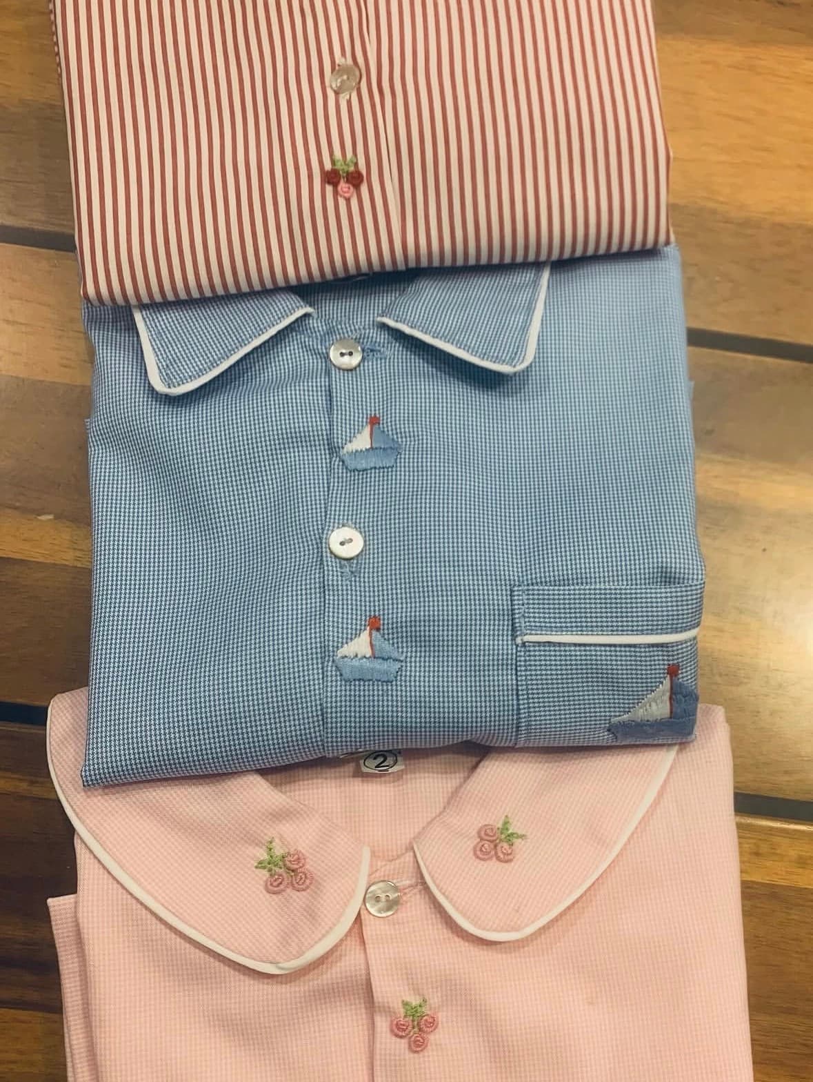 Three folded children's shirts with red stripes, blue sailboats, and pink floral embroidery on wood.