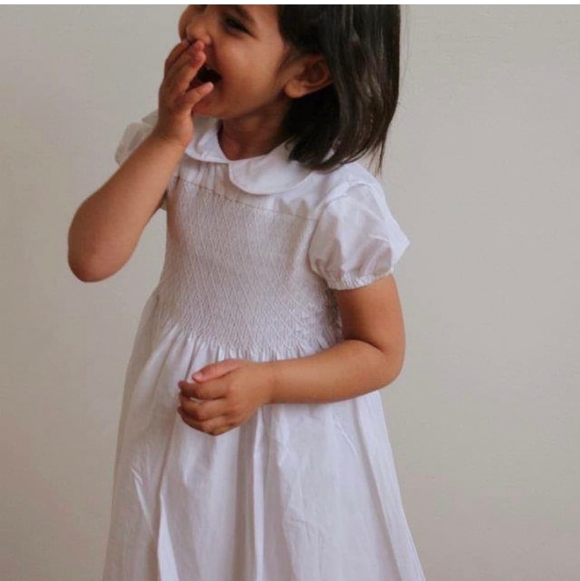 Laughing young girl in a white smocked dress with her hand over her mouth.