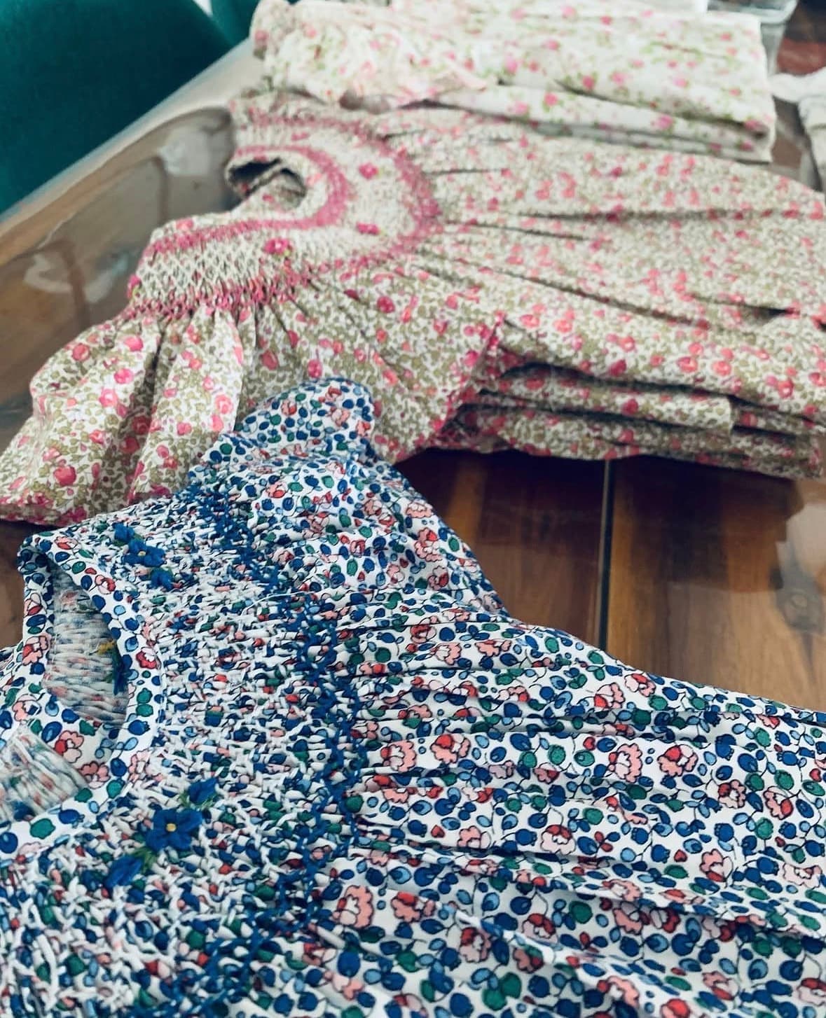 Floral print baby dresses with intricate smocked necklines folded neatly on a wooden surface.