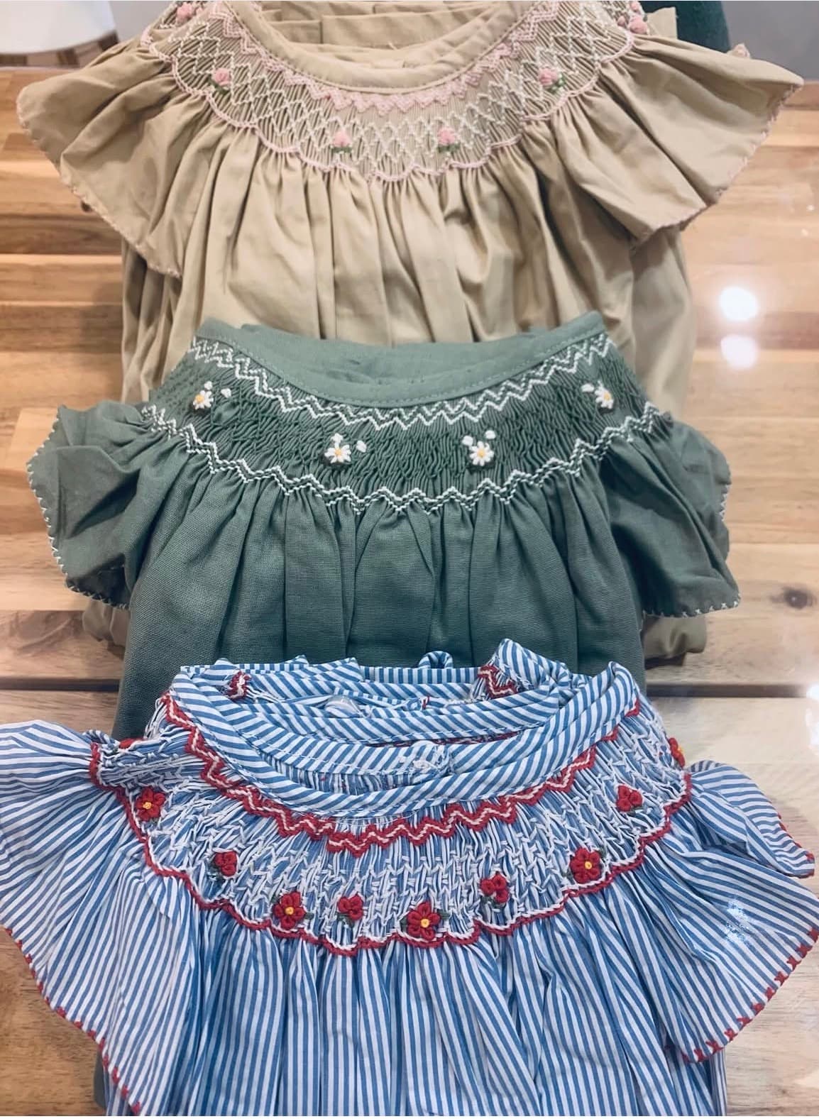 Three smocked children's dresses in beige, sage green, and blue stripes with floral embroidery.