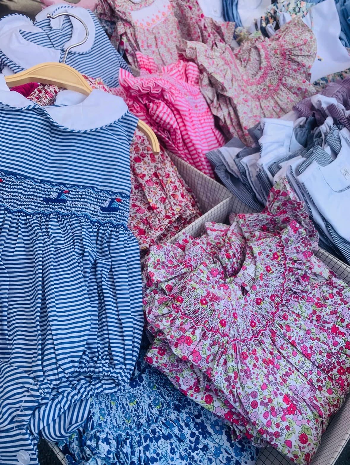 Blue striped smocked romper on a hanger next to baskets of floral children's dresses.