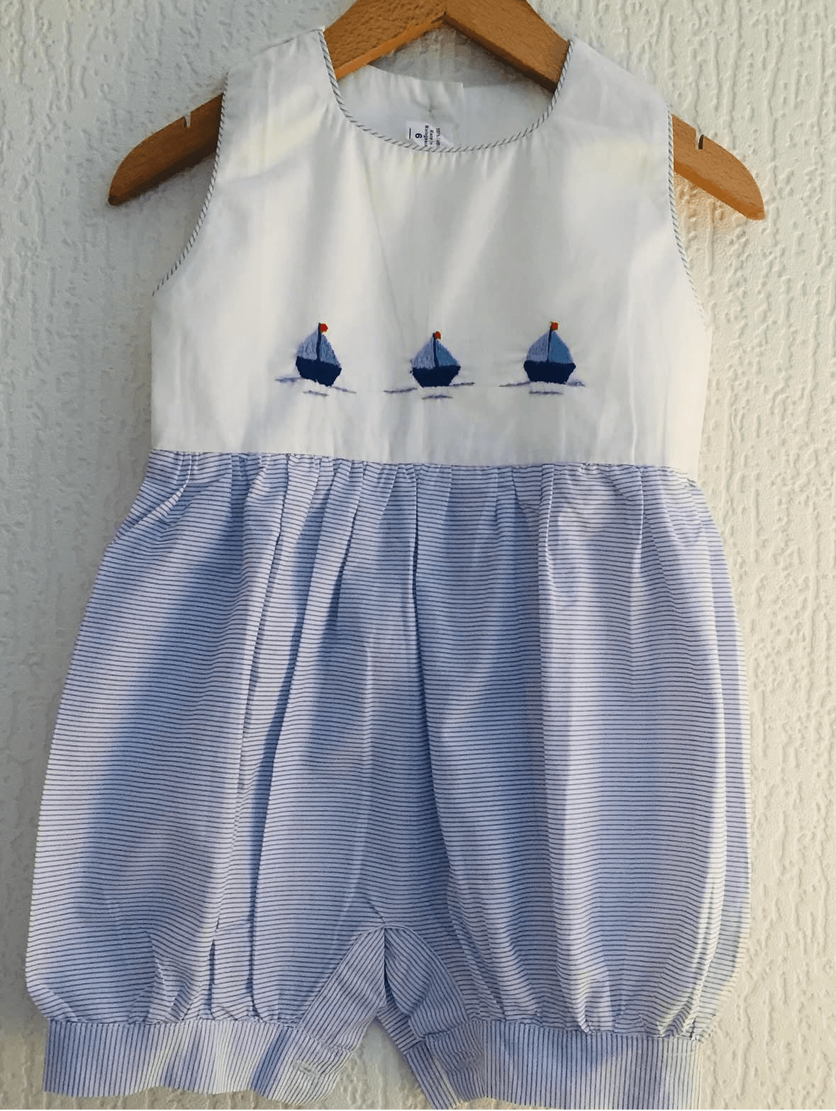 White and blue striped baby bubble romper with embroidered sailboats hanging on a wooden hanger.