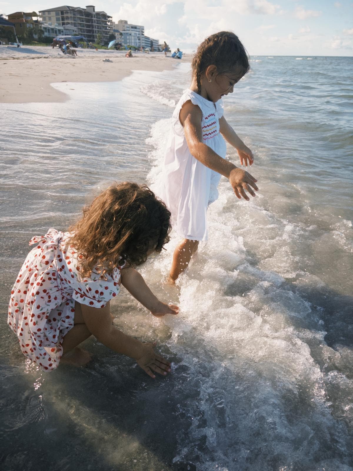 Two young girls in white dresses play in the shallow ocean waves at a sunny beach.