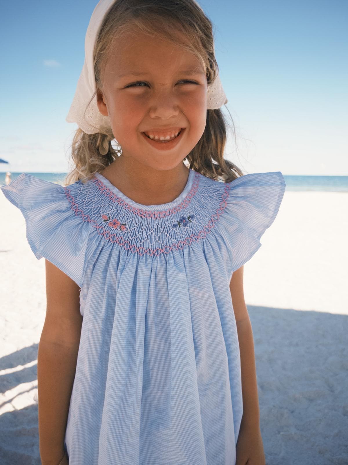 Smiling girl in a blue smocked dress and white bonnet on a sunny beach.