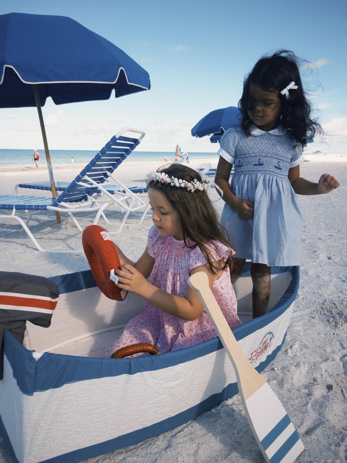 Two young girls in dresses play with a toy boat on a sunny sandy beach.