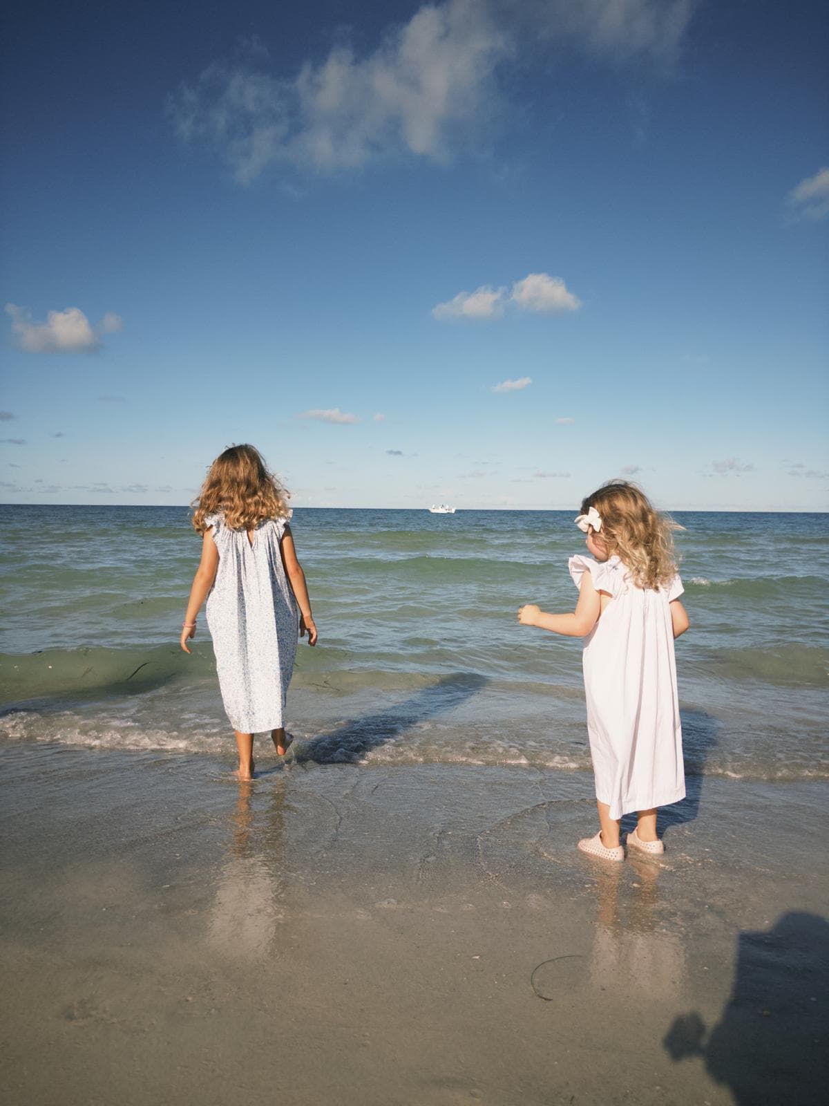 Two young girls in white dresses stand in the shallow surf, looking at the ocean.
