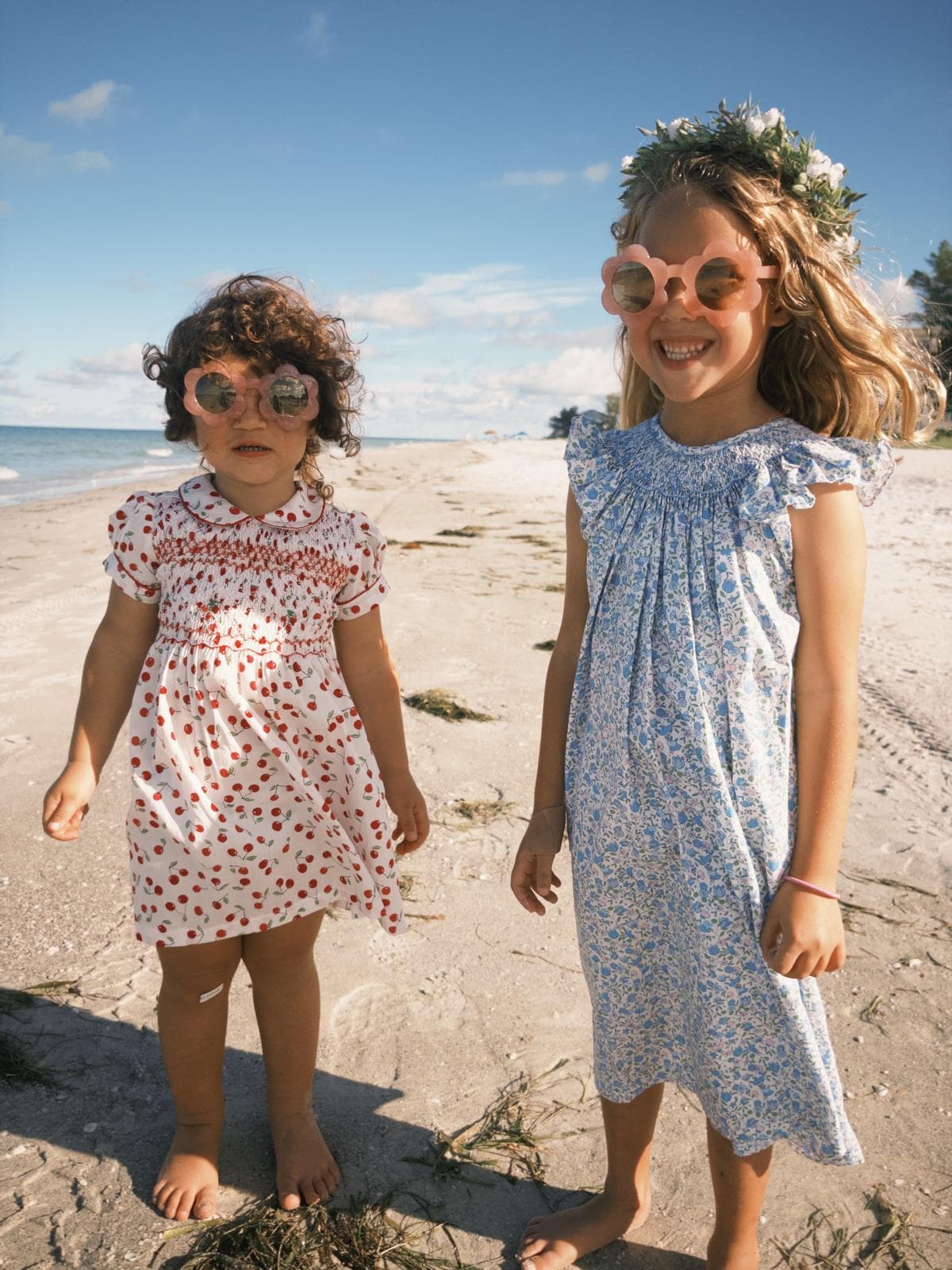 Two young girls in patterned dresses and flower-shaped sunglasses stand on a sunny sandy beach.