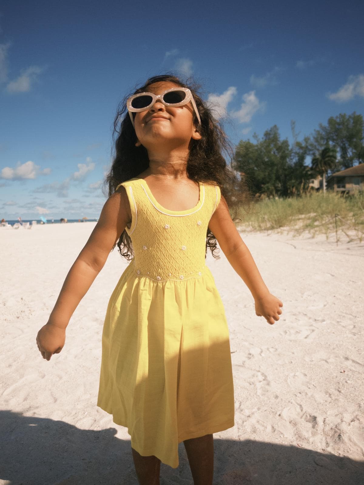 Young girl in a yellow dress and sunglasses smiles on a white sand beach.