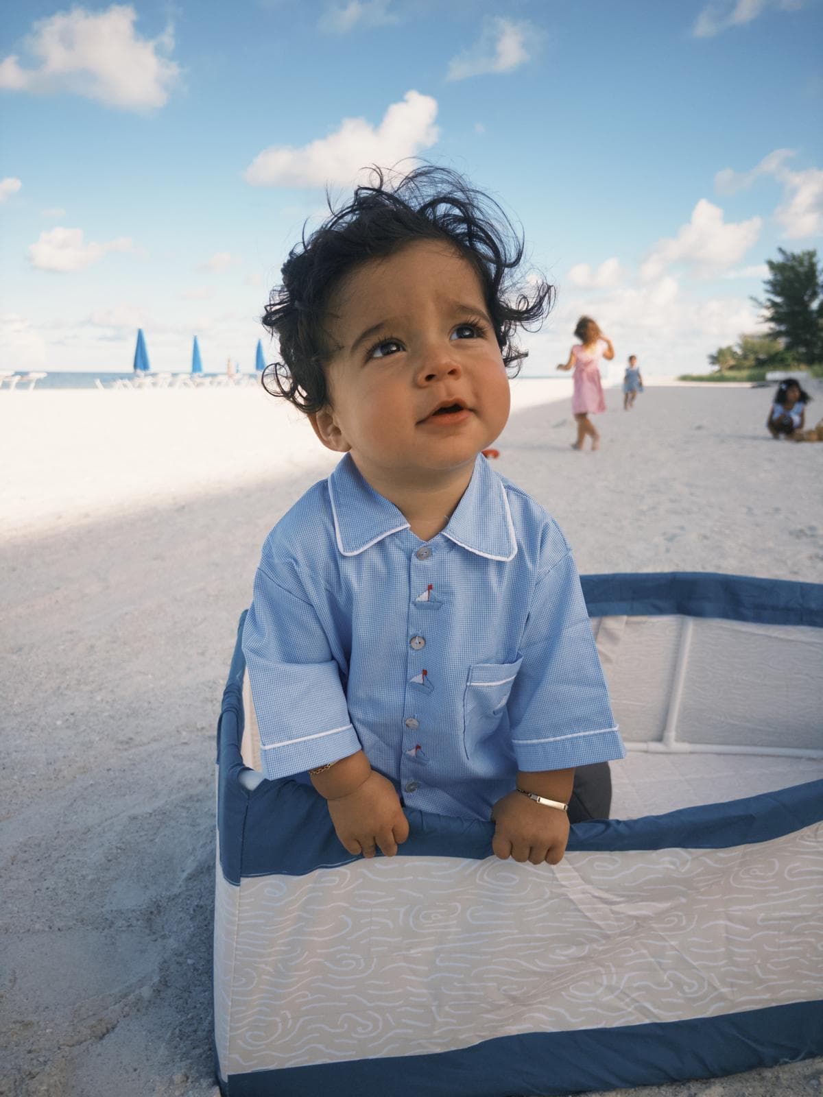 Toddler with curly hair in a blue shirt gazes upward from a beach playpen.