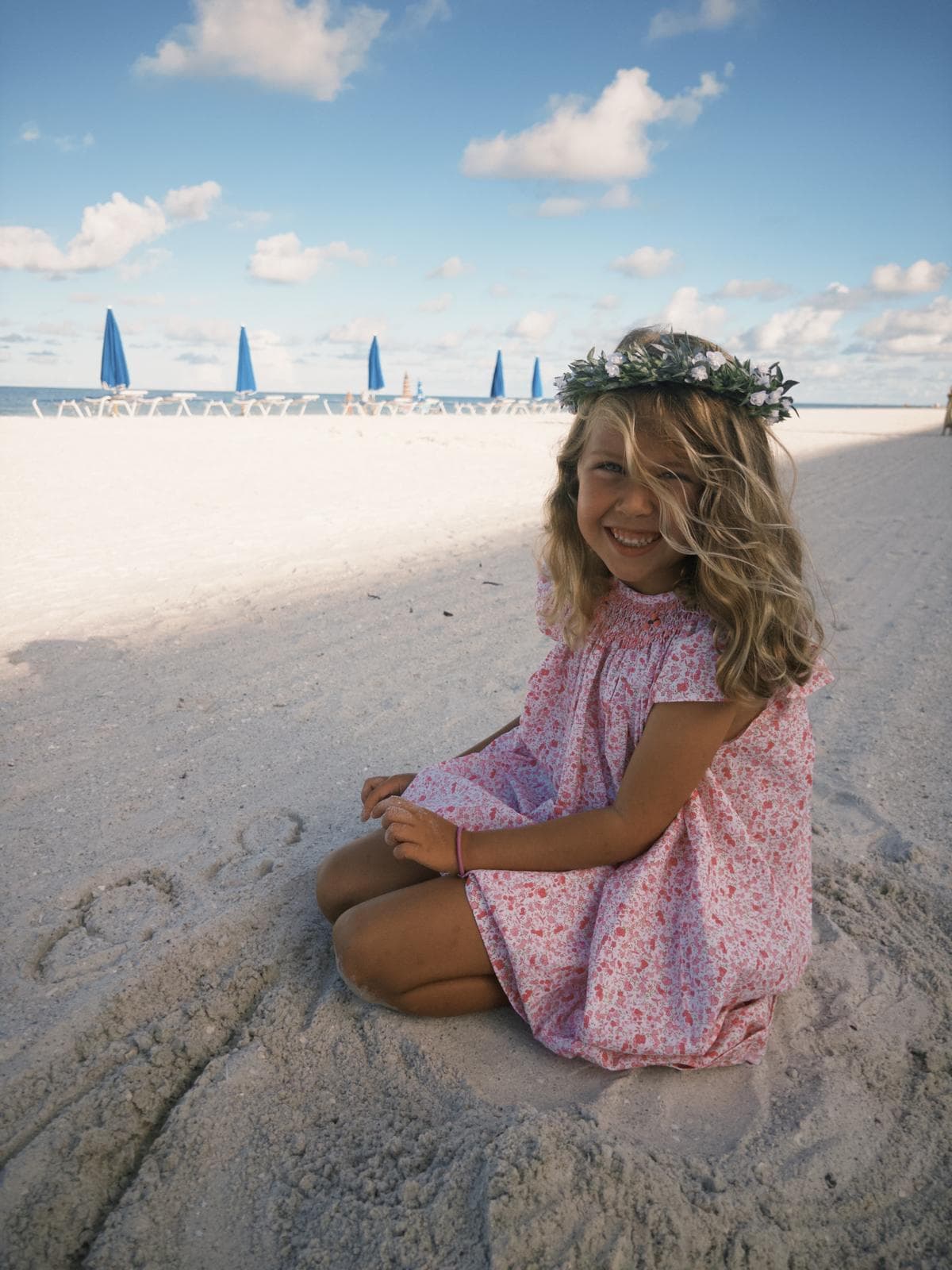 Smiling girl in a pink floral dress and flower crown sits on a white beach.
