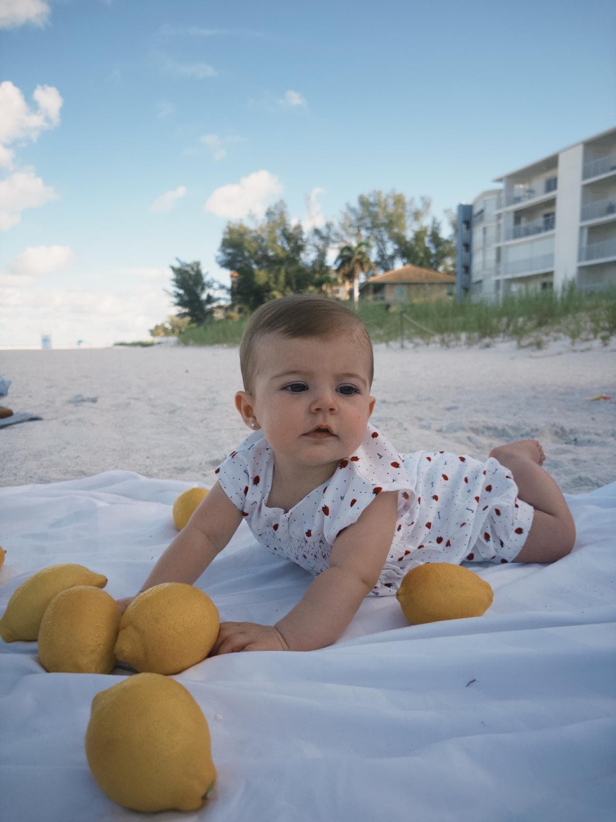 Baby in a white romper lies on a beach blanket surrounded by bright yellow lemons.